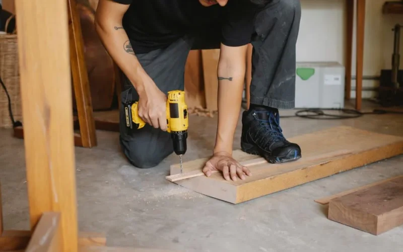 Crop anonymous male drilling piece of timber detail with screwdriver among lumber in workshop