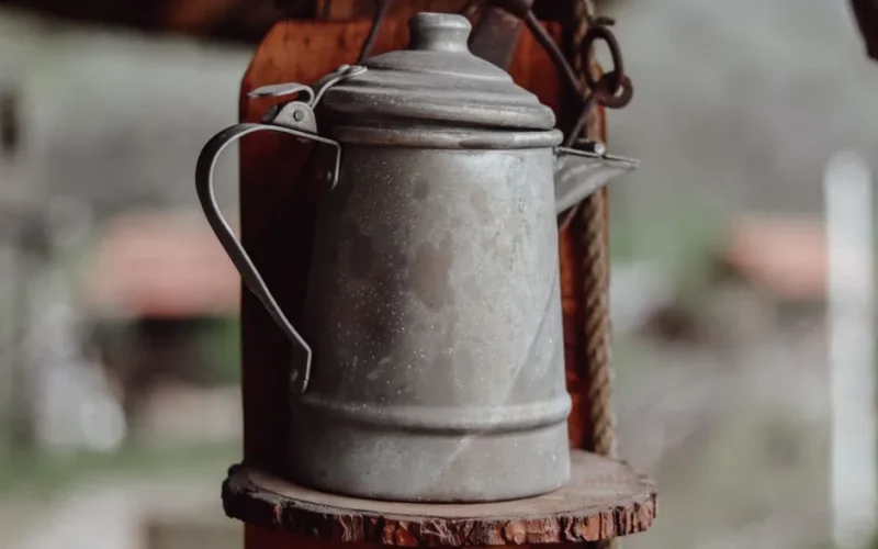 Rustic metal kettle with handle and spout displayed on a wooden shelf.