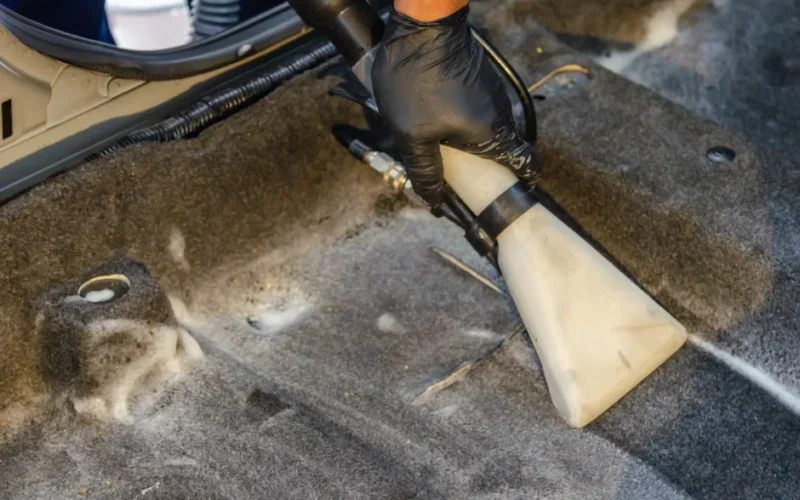 A hand in a glove using a vacuum to clean a car interior carpet, removing dirt effectively.