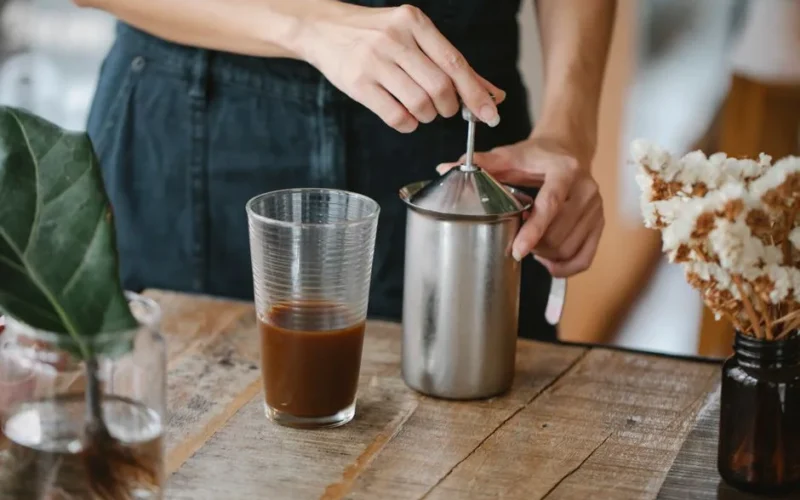 Crop anonymous female barista making froth in manual steel milk frother while preparing iced chocolate coffee in light kitchen
