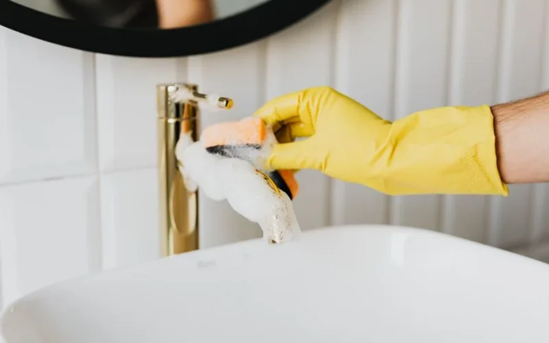 Unrecognizable man wearing yellow latex gloves washing golden faucet in bathroom while cleaning home with orange sponge and detergent