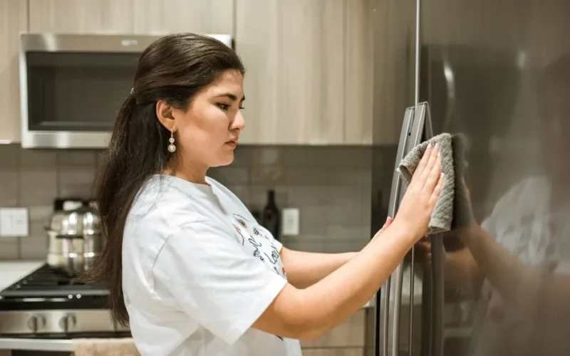 A woman cleaning a stainless steel refrigerator in a modern kitchen with a cloth.