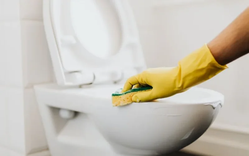 Crop anonymous person in yellow gloves cleaning white toilet rim with detergent and sponge in bathroom