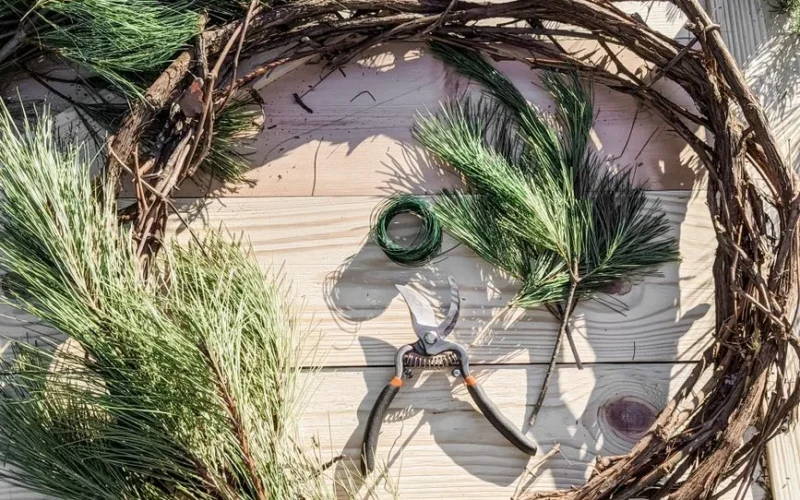 Top view of a DIY pine wreath with tools and branches on a wooden table.