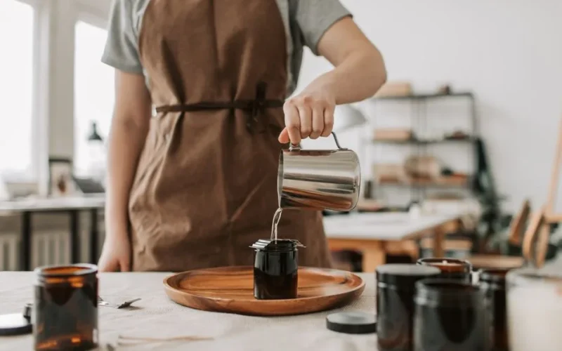 Person pouring wax into glass jars, creating handmade candles in a cozy workshop setting.