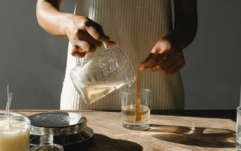 Unrecognizable African American female pouring melted wax from beaker into glass mold with wooden wick while making candles at table
