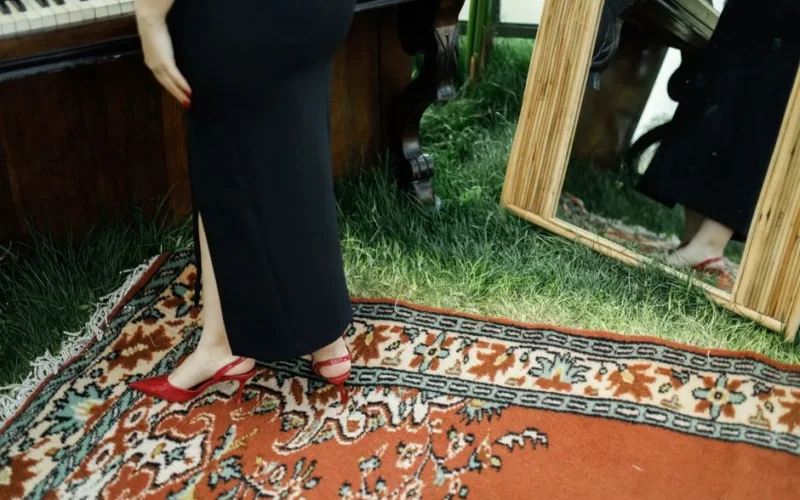 Elegant woman in black dress and red heels beside piano and mirror on patterned carpet.