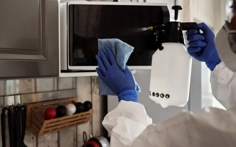 Individual in protective gear cleaning microwave using spray bottle and cloth indoors.