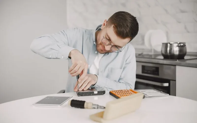 Man wearing glasses using a screwdriver to fix a device on a kitchen table.