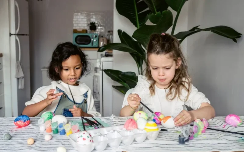 Concentrated multiracial girls painting white eggs with paintbrushes while sitting at table with paints in kitchen during Easter holiday at home