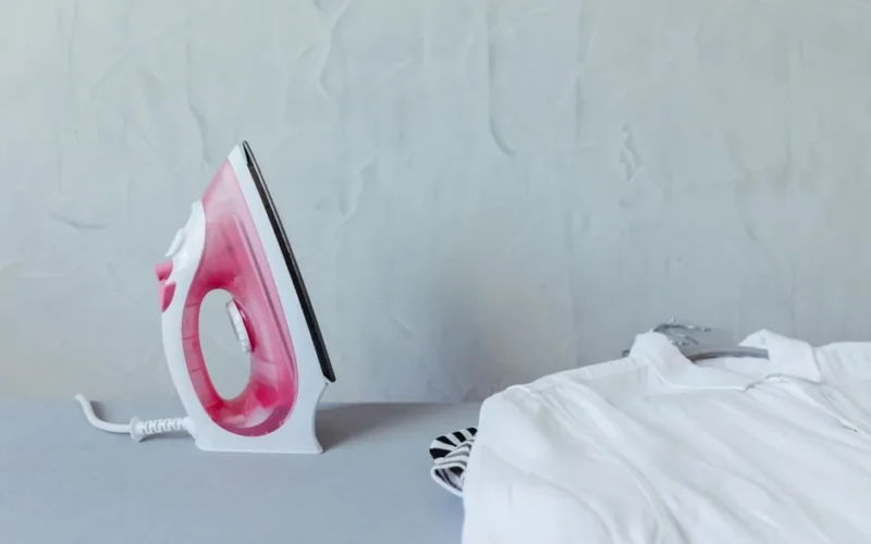 A vibrant pink iron next to a neatly folded white shirt on an ironing board against a textured wall.