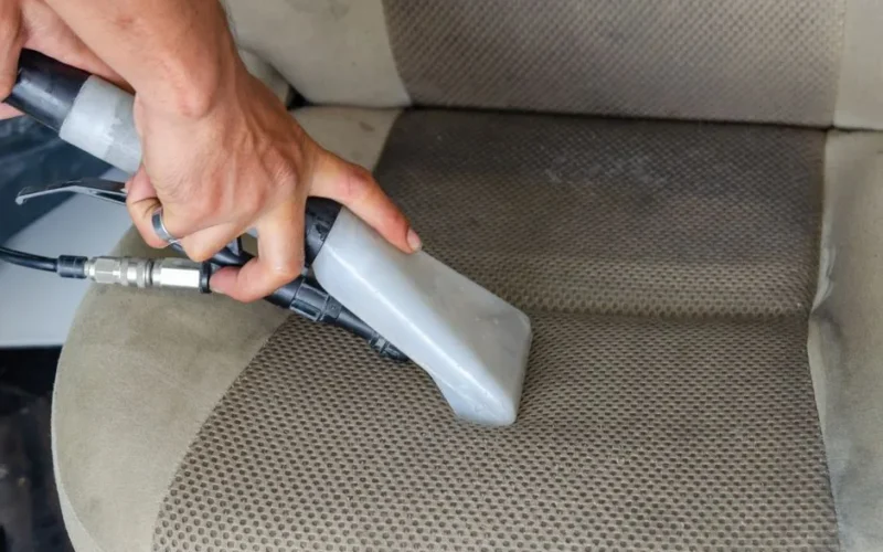 Hands using a vacuum nozzle to deeply clean a fabric car seat, removing dirt and stains.