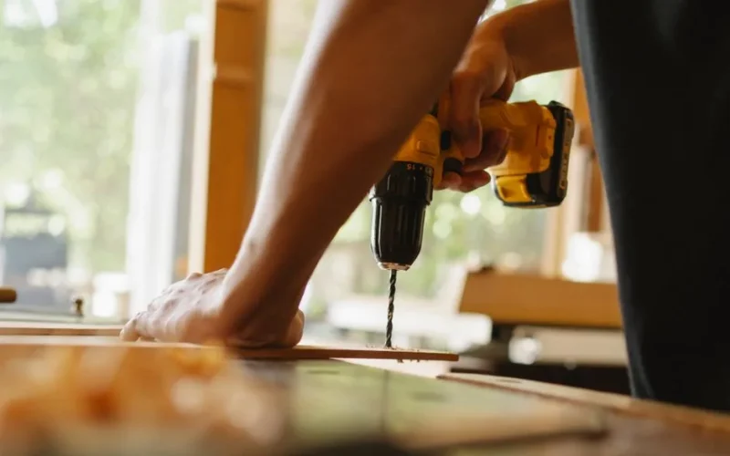 Crop anonymous male drilling timber plank with professional screwdriver on table in workshop