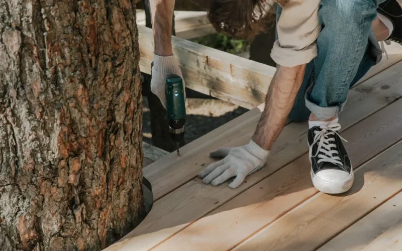 Worker drilling wooden planks on deck beside tree using power tool. DIY construction project.
