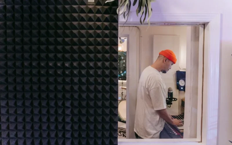 Side view of a man in a music studio with soundproof material, focused on a laptop.