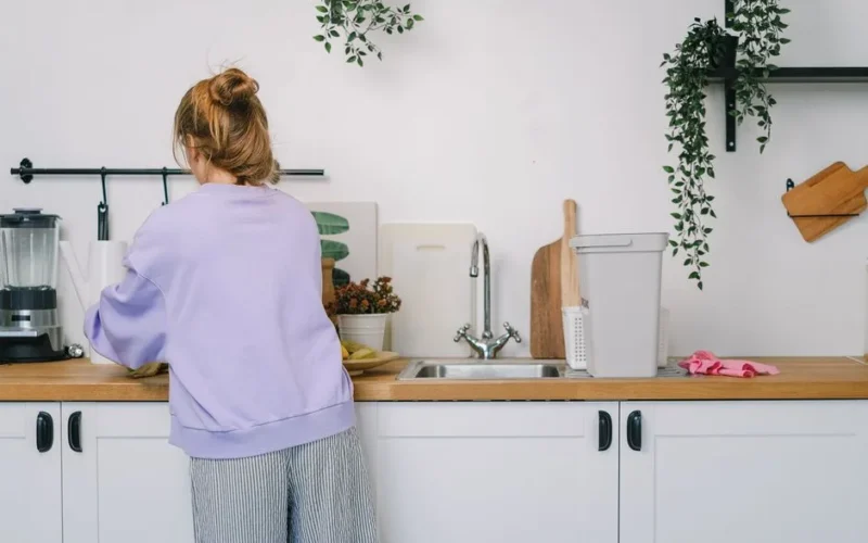 Back view of unrecognizable young female in casual clothes standing at wooden counter in light kitchen in daytime