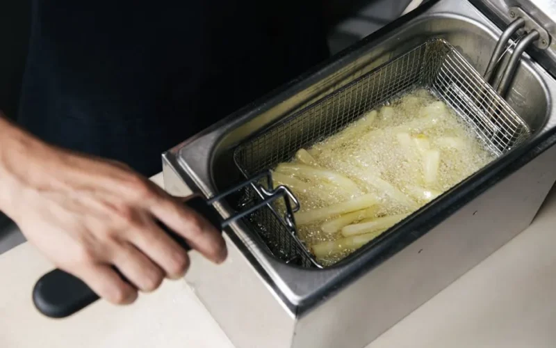A hand operates a deep fryer cooking fresh french fries in oil.