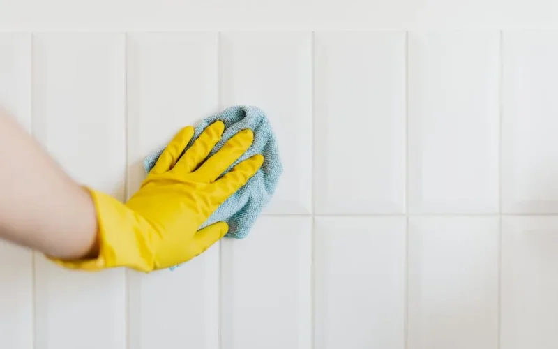 Crop anonymous housekeeper in yellow gloves cleaning white tiled wall using rag in bathroom
