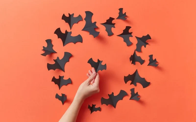 A hand arranges paper bats on a vibrant orange background, ideal for Halloween decoration concepts.
