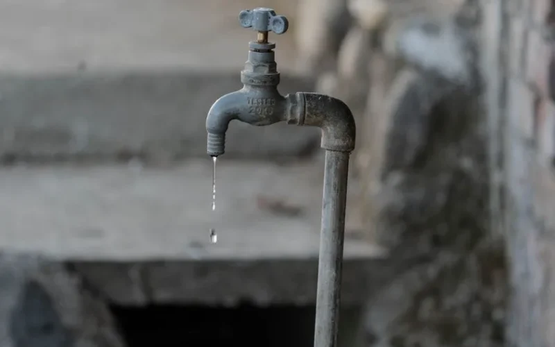 A close-up of a rustic outdoor faucet dripping water against a natural, textured background, symbolizing scarcity.