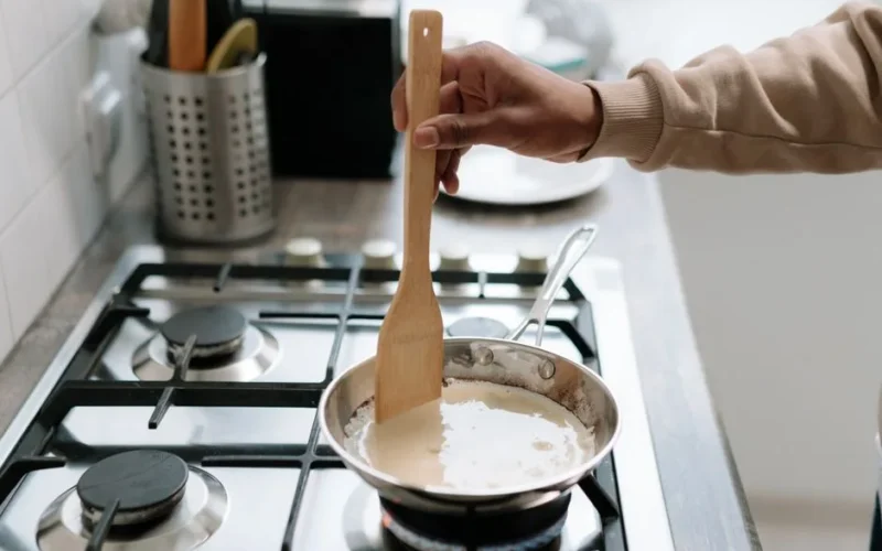 Person stirring cream sauce in a pan on a gas stove, using a wooden spatula.