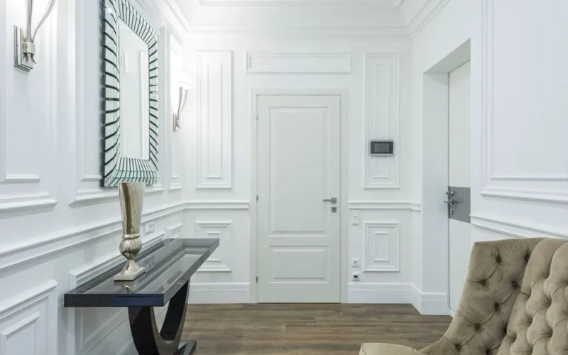 Sophisticated white interior with classic moldings and minimalist design, featuring a stylish mirrored wall and cozy seating.