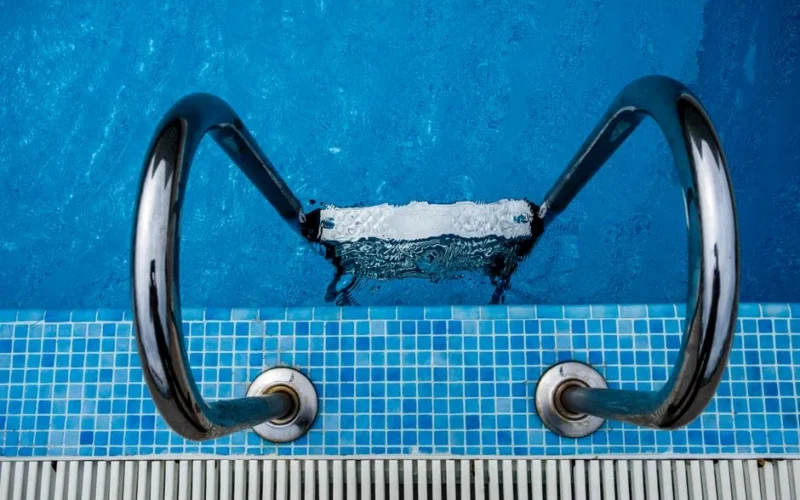 Aerial view of a stainless steel ladder entering a sparkling blue swimming pool with tiled edges.