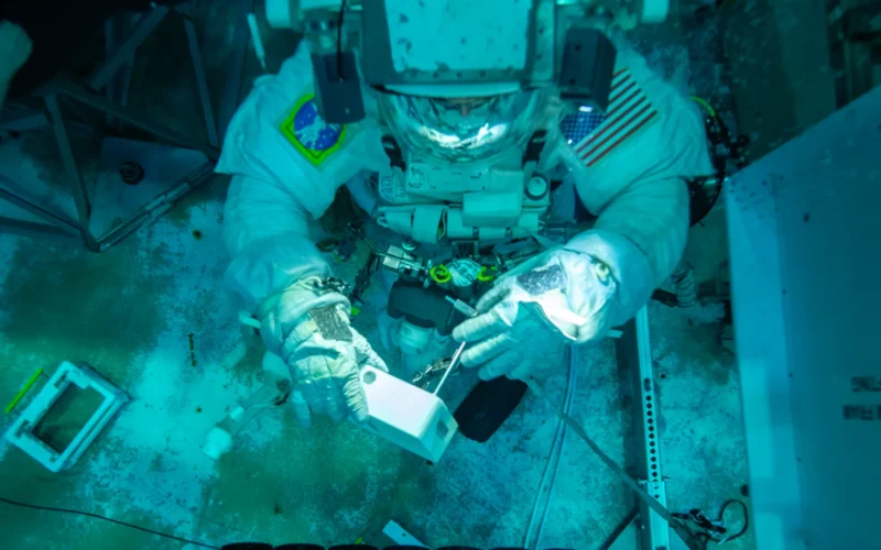 Astronaut Don Pettit removes a patch from the practice NICER caddy during a training exercise at the NBL at NASA’s Johnson Space Center in Houston on May 16, 2024. Credit: NASA/NBL Dive Team Alt text: An astronaut in a pool. Image description: A man near the bottom of a pool wears a spacesuit. The camera looks down on him from above. He hovers in the top half of the image. His helmet light illuminates his hands. One hand holds a white plastic box. The other hand holds a silver T-shaped tool, which he pokes into the box. In the bottom half of the image is a mockup of the NICER instrument. It’s a hollow square. The side facing Pettit is covered in rows of black circles. Each circle is segmented internally into six pieces, like a pie. The concrete pool floor is visible beneath the man and model.