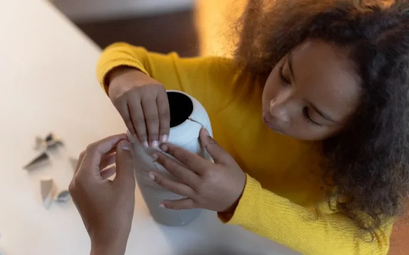 A young girl repairing a ceramic vase with an adult's assistance indoors.