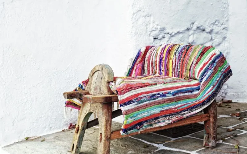 A vibrant, multicolored rug draped over a rustic wooden chair against a white stucco wall.