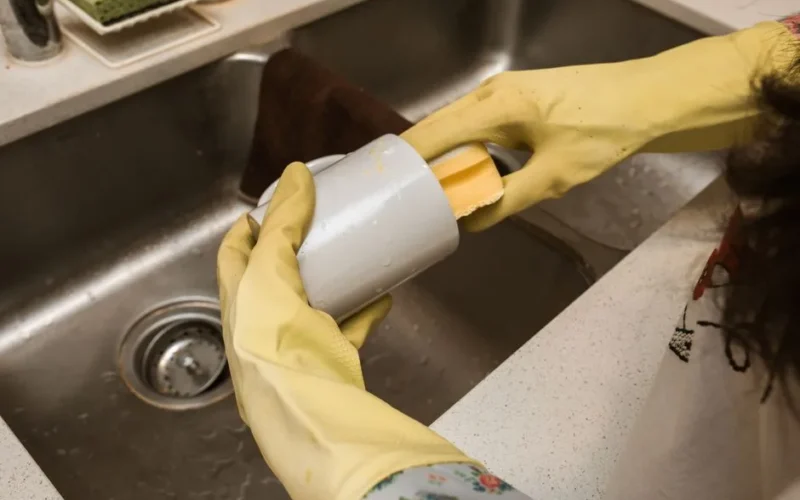 Close-up of a person washing a mug with yellow rubber gloves and sponge in a kitchen sink.