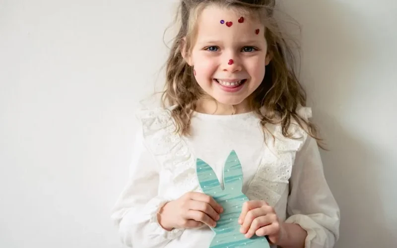 Cheerful preschool girl with heart shaped stickers on face looking at camera while standing on white background with Easter bunny decoration in hands