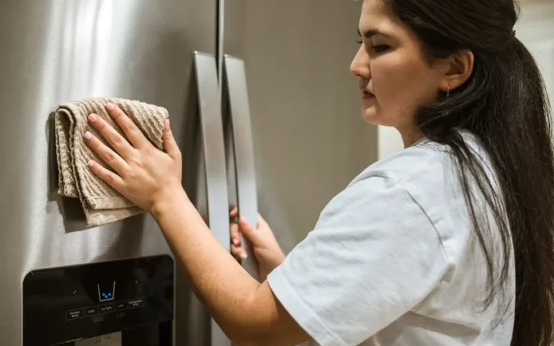 A woman wipes a refrigerator with a cloth, focusing on maintaining cleanliness in a kitchen.