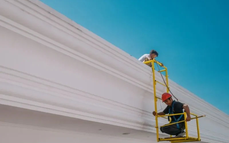 Two construction workers on a platform working on a white building under a clear sky.