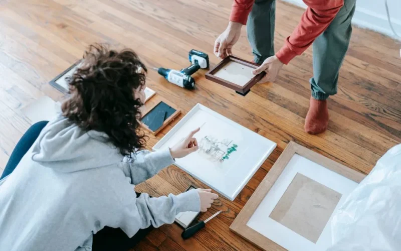 A couple organizing picture frames on a wooden floor, preparing for interior decoration or renovation.