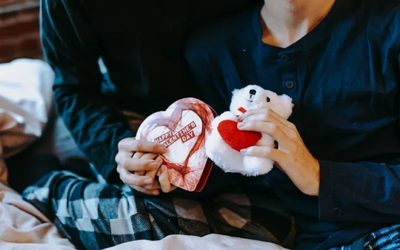 A cozy indoor scene of adults exchanging Valentine's gifts, featuring a teddy bear and heart-shaped box.