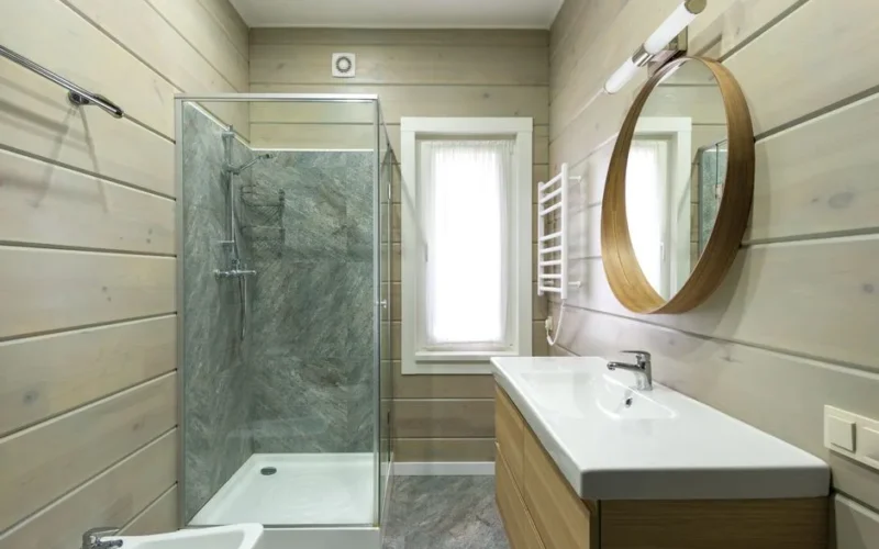 Contemporary bathroom design with round mirror hanging above sink and shower cabin near window and paneled walls