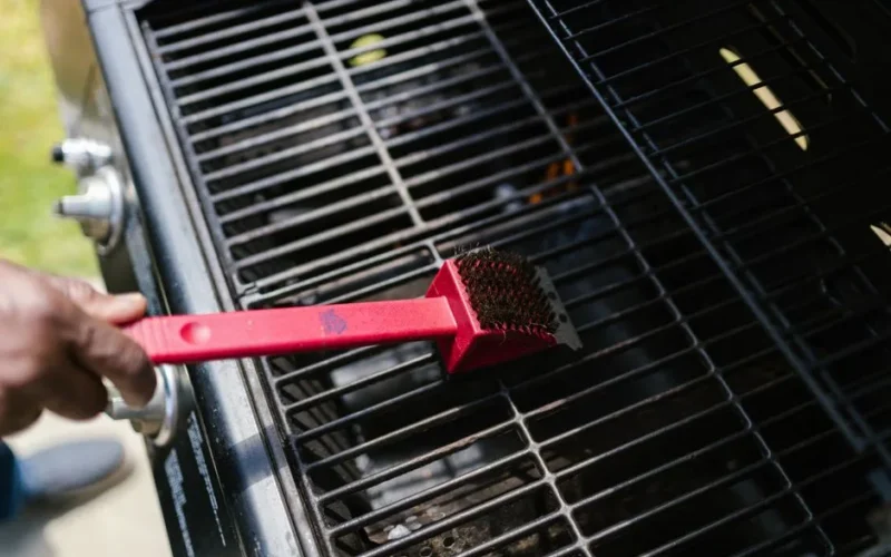 A person cleaning a barbecue grill with a red brush in daylight. Ideal for BBQ enthusiasts.