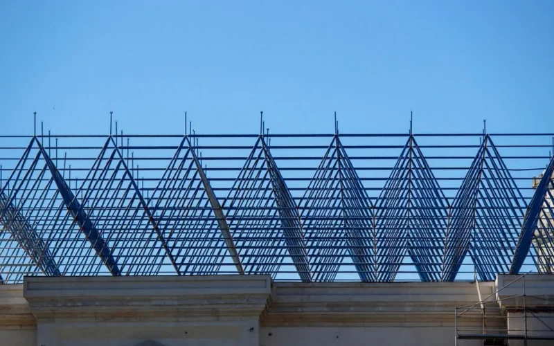 A geometric steel frame of a roof under construction on a building, set against a clear blue sky.