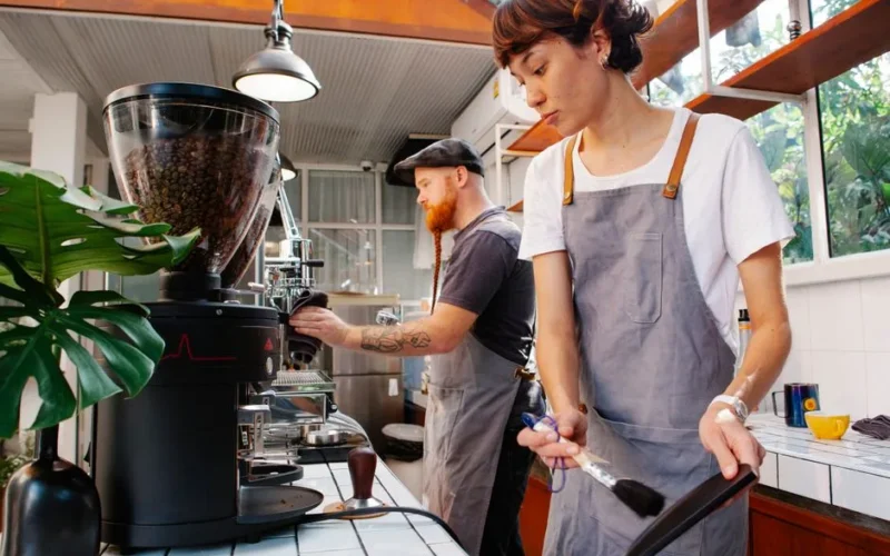 Crop female cafeteria employee with brush near bearded male partner against coffee machine working in daylight