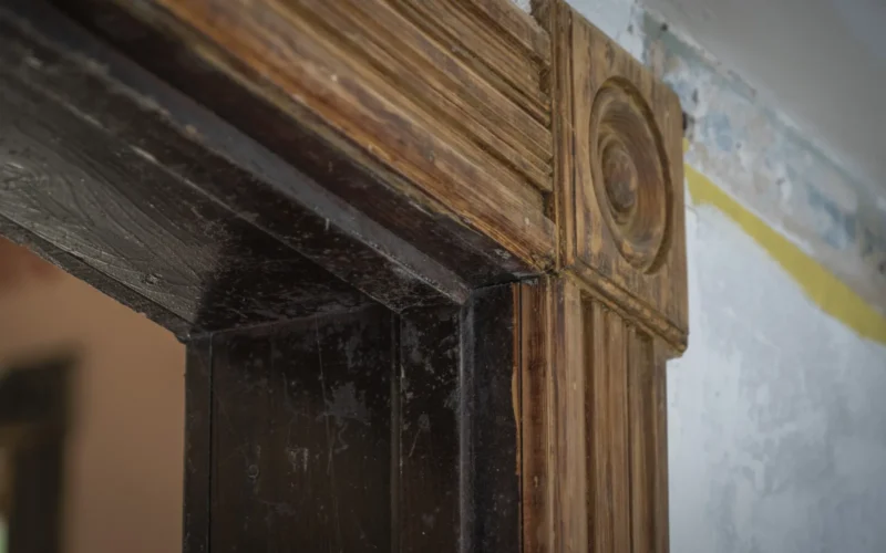 The decorative corner of a doorframe, showing dark finish inside and light finish against the wall. A contrast between the old, dark finish (inside the door frame) and the bare wood color of the trim inside the church.