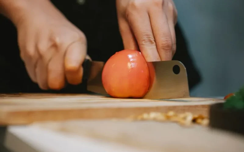 Crop unrecognizable chef slicing ripe wet red tomato with knife on wooden cutting board for cooking healthy dish