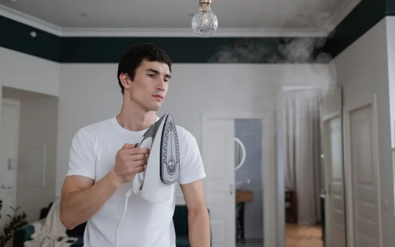 A man uses a steam iron on a white shirt indoors, highlighting daily household chores.